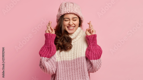 Hopeful optimistic young lovely woman with glad expression, makes wish to accomplish goals, raises hands with crossed fingers, wears oversized winter jumper and hat, isolated over pink background. photo