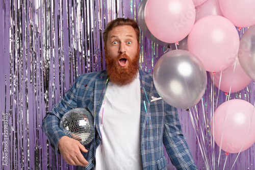 Emotional stunned red haired bearded man carries glitter disco ball and bunch of air balloons, comes on costume party, surprised to notice drunk friend, poses over lilac sparkling decorated wall. photo