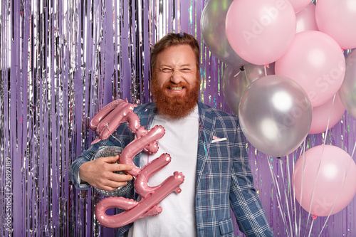 Photo of optimistic emotional red haired man with thick beard, laughs happily, dressed in trim jacket, holds balloons and disco ball, comes on theme party, makes photo with decor accessories