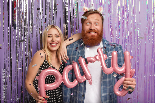 Redhead birthday man in festive mood, glad to celebrate his holiday in company of noisy friends, poses for making photo with girlfriend, hold pink letter shaped balloons, throw big blow out. photo