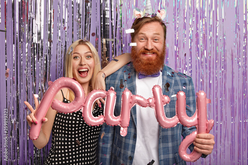 Two young male and female enjoy social gathering, invited to big party, hold balloons, come on receiption, wear formal clothes, sparkling tinsel curtain in background, have fun and joy together photo