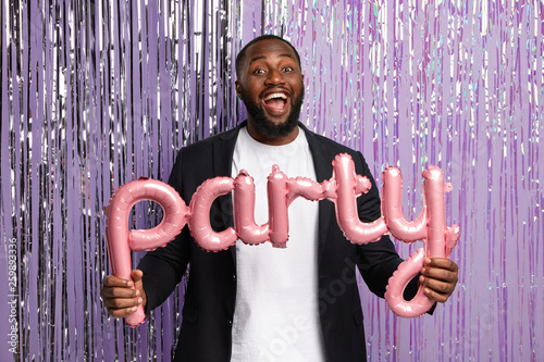Happy dark skinned man in elegant suit, throws party, holds pink balloon letters, has joyful look, enjoys entertainment with friends loud music stands against purple background with sparkle decoration photo