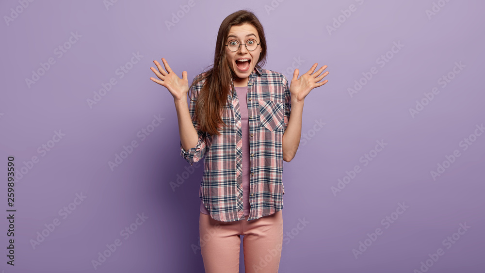 Photo of joyful emotional woman spreads hands, exclaims from happiness, wears checkered shirt, looks with eyes full of positive emotions, isolated over purple background, screams in surprise Photo of joyful emotional woman spreads hands, exclaims from happiness, wears checkered shirt, looks with eyes full of positive emotions, isolated over purple background, screams in surprise