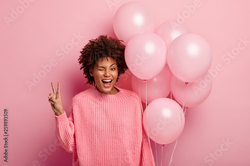 Smiling joyful dark skinned female model blinks eye, shows peace gesture, dressed casually, carries bunch of festive balloons, expresses happiness, isolated over pink background. Studio shot © wayhome.studio 