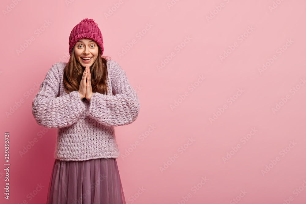 Indoor shot of pleased happy female keeps palms pressed together, prays and hopes for better, has glad facial expression, dressed in warm fashionable clothes, stands over pink wall with free space Indoor shot of pleased happy female keeps palms pressed together, prays and hopes for better, has glad facial expression, dressed in warm fashionable clothes, stands over pink wall with free space