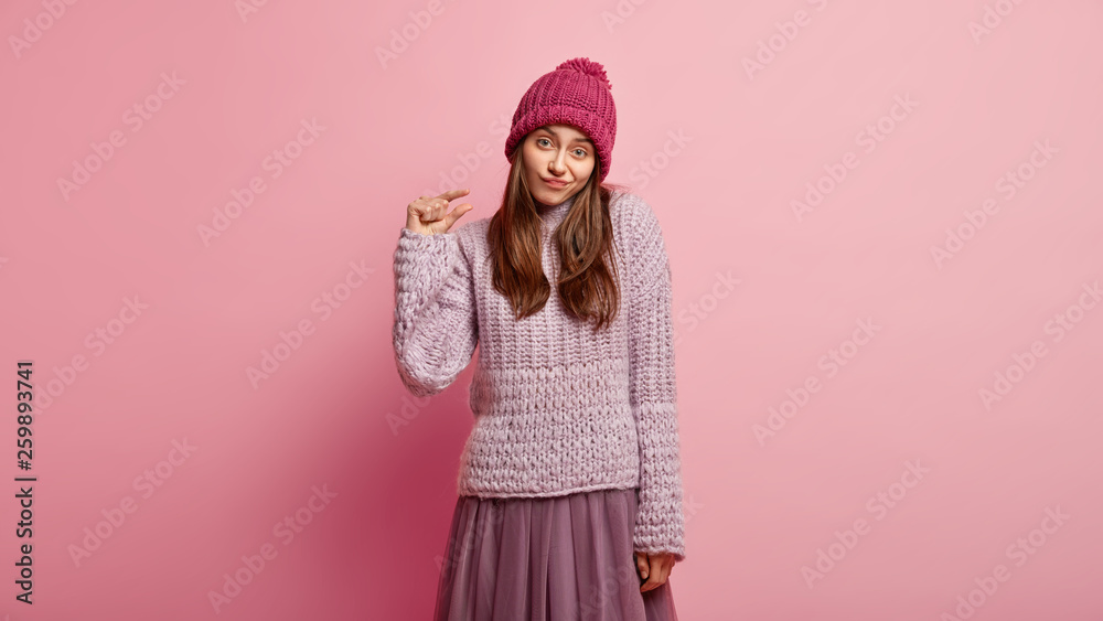 Unimpressed attractive young woman shows something tiny, demonstates little sign, wears pink headgear and winter sweater, makes hand gesture, offers to drink small amount of coffee. Size concept Unimpressed attractive young woman shows something tiny, demonstates little sign, wears pink headgear and winter sweater, makes hand gesture, offers to drink small amount of coffee. Size concept