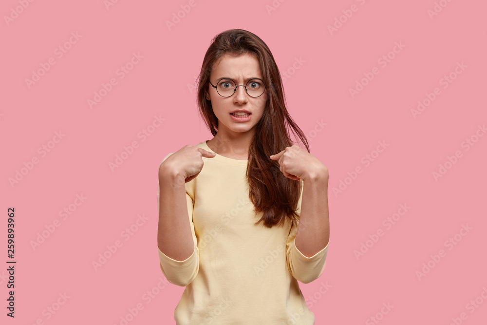 Photo of embarrassed lovely woman points at herself, has annoyed facial expression, wears transparent glasses and jumper, reacts emotionally on being blamed, stands indoor over pink background. Photo of embarrassed lovely woman points at herself, has annoyed facial expression, wears transparent glasses and jumper, reacts emotionally on being blamed, stands indoor over pink background.