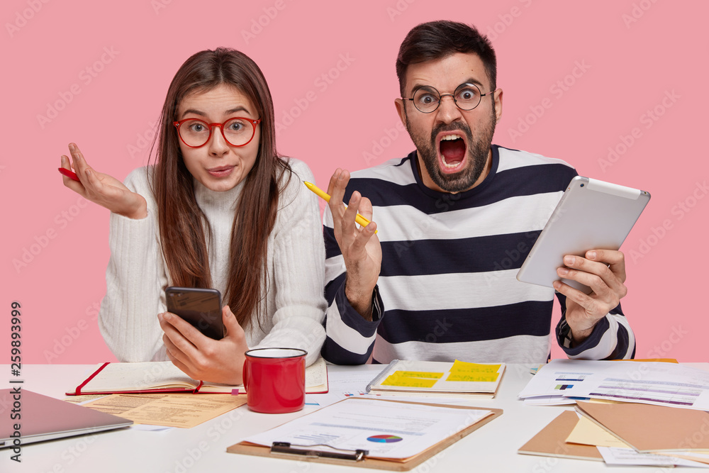 Horizontal shot of confused female and annoyed man discuss graphs, work on university project, use modern technologies, do homework together, isolated over pink background. Youth and work concept Horizontal shot of confused female and annoyed man discuss graphs, work on university project, use modern technologies, do homework together, isolated over pink background. Youth and work concept