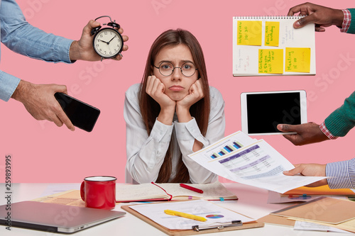 Sad female workaholic keeps hands under chin, busy making project work, studies papers, wears elegant white shirt, sits at desktop, unknown people stretch hands with notes, alarm clock, smartphone
