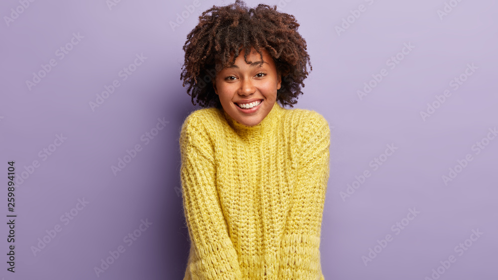 Horizontal shot of attractive dark skinned female has sincere smile, feels optimistic and glad, being in good mood, dressed in yellow jumper, poses against lilac studio wall. People and emotions Horizontal shot of attractive dark skinned female has sincere smile, feels optimistic and glad, being in good mood, dressed in yellow jumper, poses against lilac studio wall. People and emotions