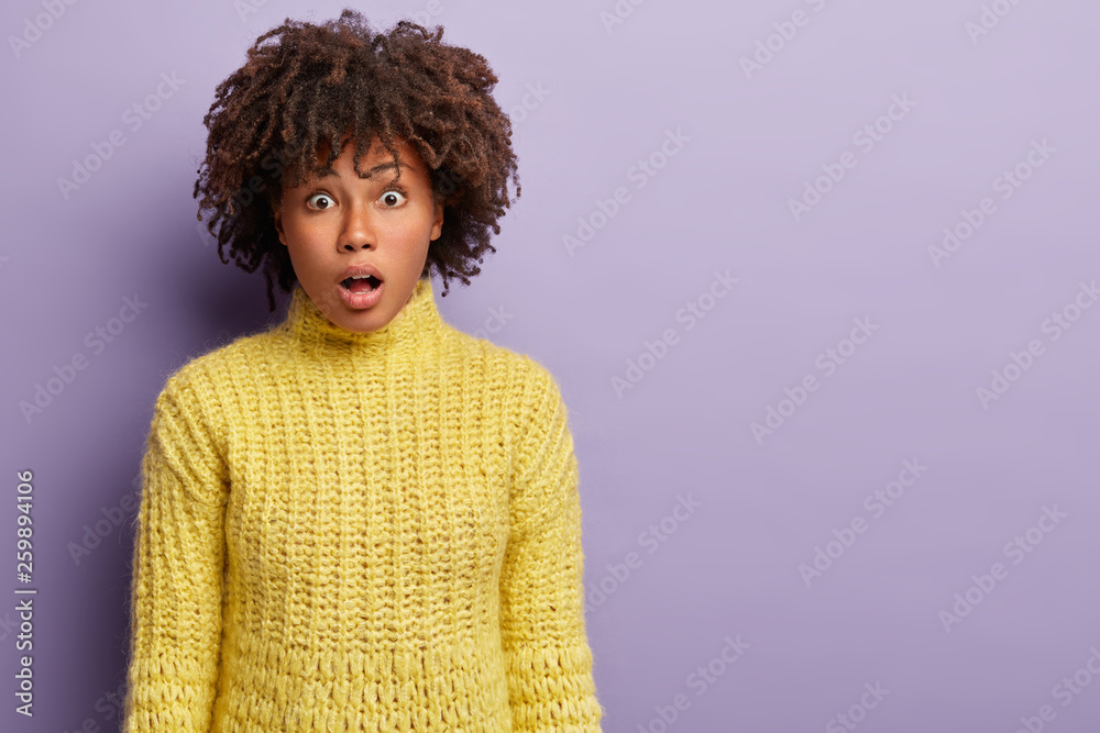 People, facial expressions concept. Stupefied dark skinned female model opens mouth, stares at camera, expresses shock, hears bad latest news, wears yellow clothes, isolated over purple wall People, facial expressions concept. Stupefied dark skinned female model opens mouth, stares at camera, expresses shock, hears bad latest news, wears yellow clothes, isolated over purple wall