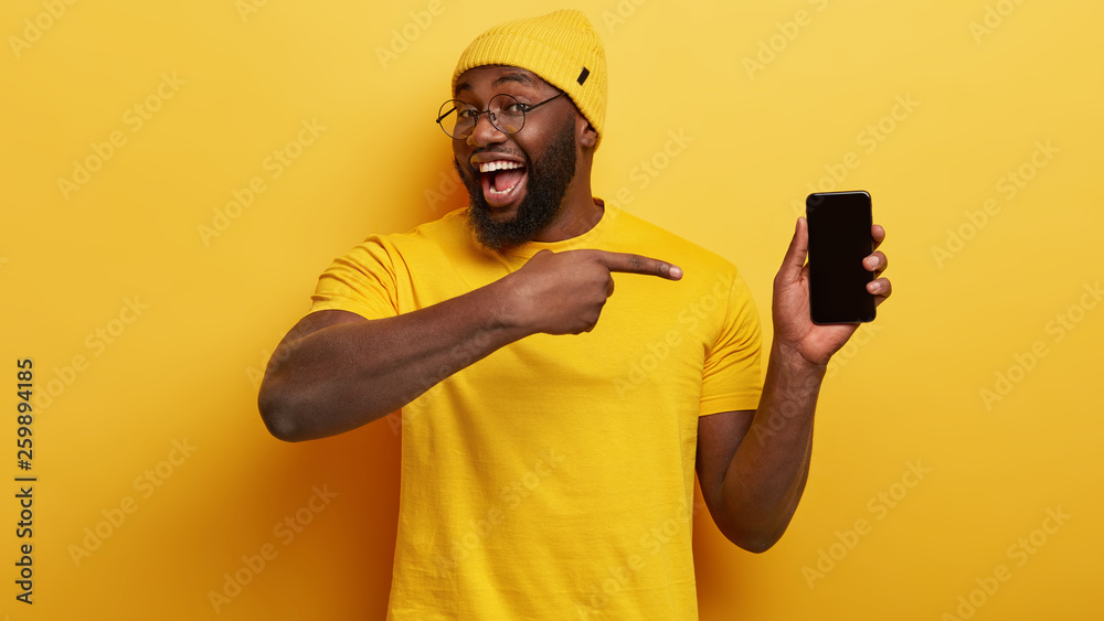 Delighted dark skinned male seller of modern devices, points at mobile phone with mock up black screen suggests buying gadget for low price wears yellow fashionable clothes in one tone with background Delighted dark skinned male seller of modern devices, points at mobile phone with mock up black screen suggests buying gadget for low price wears yellow fashionable clothes in one tone with background