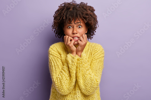 Image of nervous dark skinned female bites finger nails from depression  worries because of hurt feelings and seperation with boyfriend  has Afro haircut  wears yellow jumper  poses indoor alone