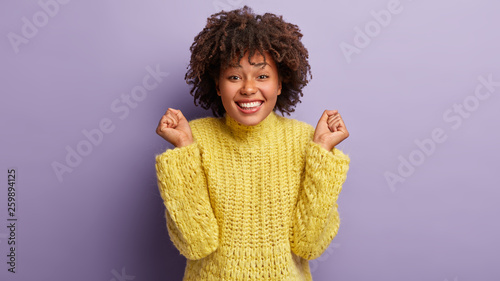 Optimistic dark skinned girl clenches fists in triumph, celebrates successfully passed entrance exam, smiles gladfully, has Afro haircut, dressed casually, isolated over lilac wall. People and victory