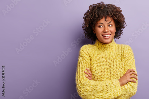 Good looking mixed race female with curly dark hair, grins at camera, shows white teeth, keeps arms folded, focused aside, wears yellow clothes, dreams about good holidays, isolated on purple