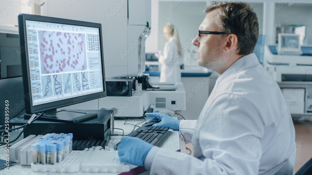 Male Scientist Sits at His Workplace in Laboratory, Uses Personal Computer. Screen Shows Analyze of DNA. I the Background Genetics Research Centre with Innovative Equipment. Male Scientist Sits at His Workplace in Laboratory, Uses Personal Computer. Screen Shows Analyze of DNA. I the Background Genetics Research Centre with Innovative Equipment.