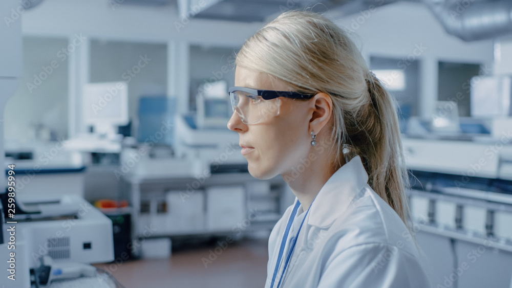 Fototapeta premium Portrait Shot of Female Scientist Sits at His Workplace in Laboratory, Uses Personal Computer. In the Background Genetics Research Centre with Innovative Equipment.