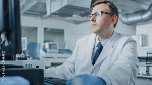 Male Research Scientist Sits at His Workplace in Laboratory, Uses Personal Computer. I the Background Genetics, Pharmaceutical Research Centre with Innovative Equipment.