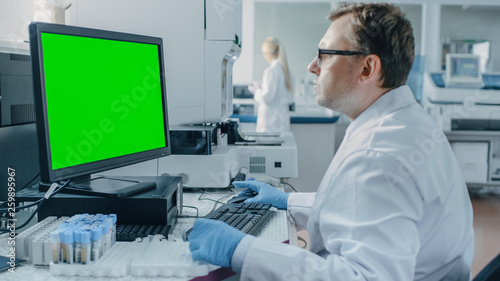 Male Research Scientist Sits at His Workplace in Laboratory, Uses Green Mock-up Screen Personal Computer. I the Background Genetics, Pharmaceutical Research Centre with Innovative Equipment.