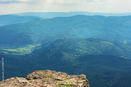 beautiful summer mountain landscape. stunning view from the edge of a hill with huge rock. grassy slopes. ridge and valley in the far distance. sunny weather with cloudy sky at high noon
