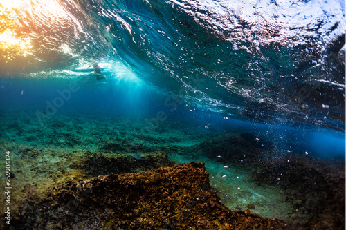 Underwater view of the ocean wave breaking over the shallow reef with sharp stones. Surfer floats on the background