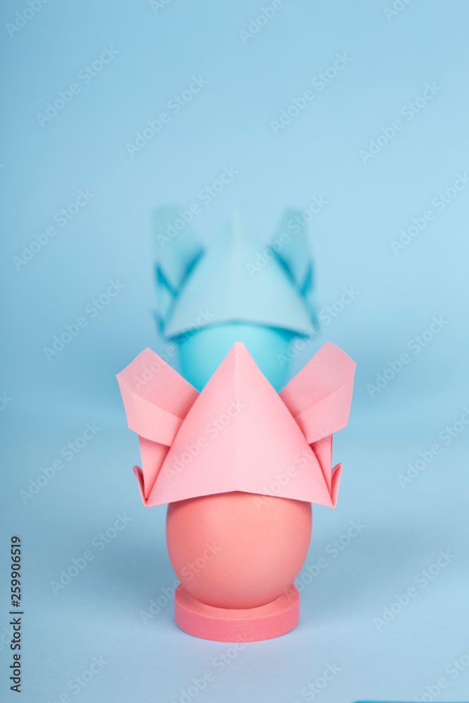 Happy Easter holiday concept dyed chicken pink and blue eggs in a bunny origami hats with ears on blue paper background with copy space. COVID-19 easter concept Happy Easter holiday concept dyed chicken pink and blue eggs in a bunny origami hats with ears on blue paper background with copy space. COVID-19 easter concept