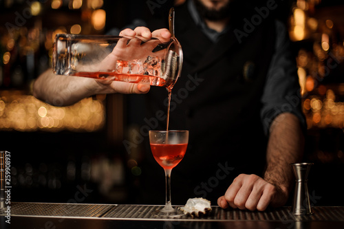 Bartender pouring a delicious red cocktail from the measuring glass cup through the strainer