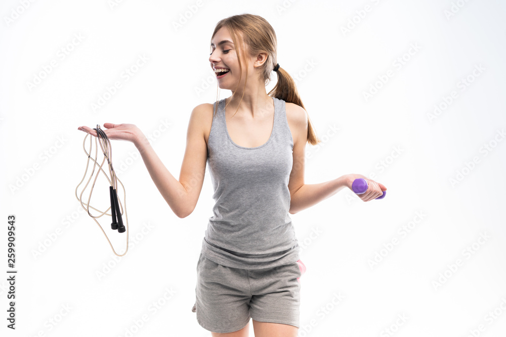 Fit happy woman holding jump rope and dumbbells looking at camera over white background. Fit happy woman holding jump rope and dumbbells looking at camera over white background.