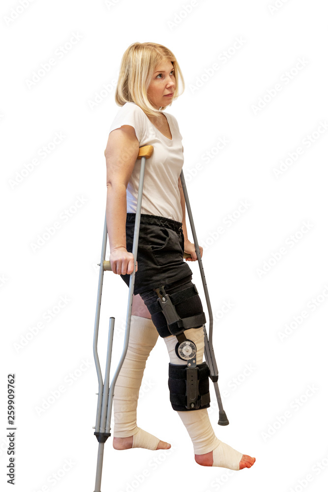 Young woman after knee surgery. On crutches, in an orthosis and with bandaged legs. Isolated on a white background. Vertical.