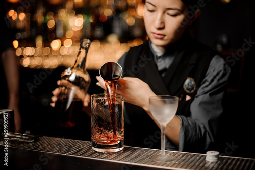 Bartender girl pouring a delicious brown cocktail from the steel jigger