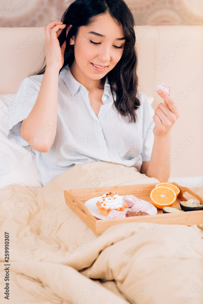 Beautiful awesome dark haired woman having tasty breakfast in bed at her cozy bedroom. Side view photo of young girl in blue striped pajamas eating sweets.