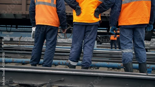 Railway workers are standing and looking at a passing train. Legs close-up. Worker in orange workwear. In the background traveling freight cars