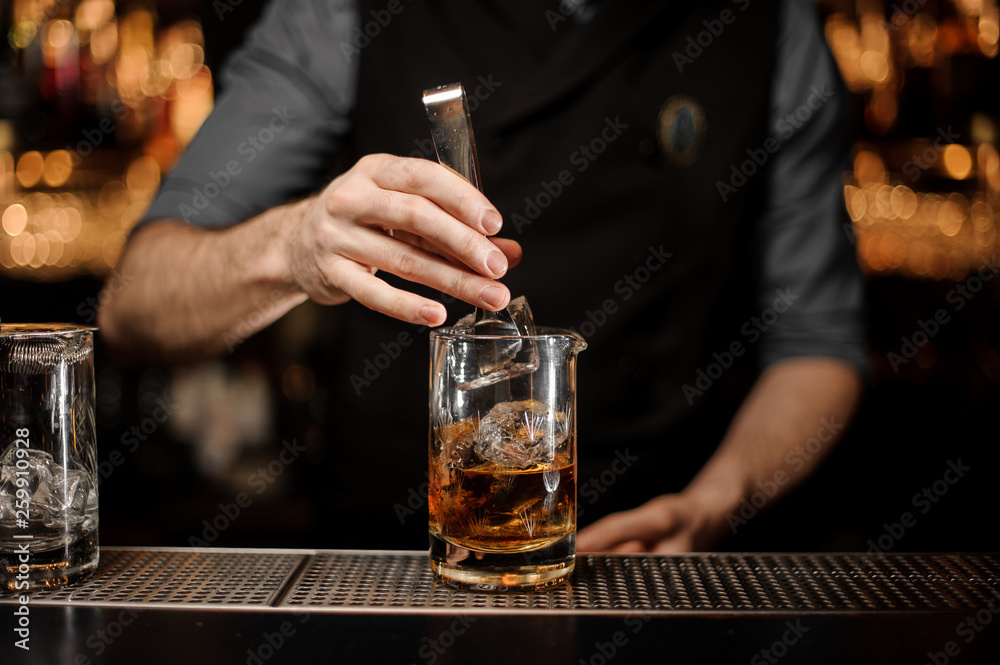 Bartender adding a big ice cube to the measuring glass cup with a brown cocktail