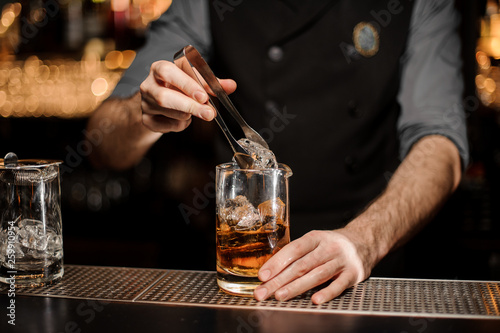 Professional bartender adding a big ice cube to the measuring glass cup with a brown cocktail