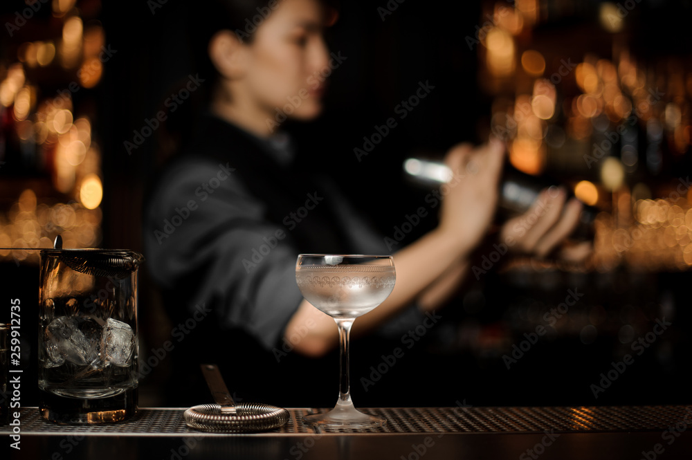 Cocktail glass on the blurred foreground of bartender girl holding a steel shaker