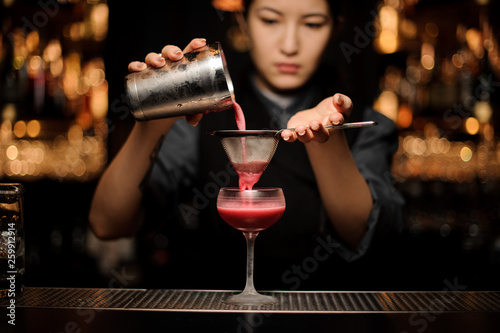 Professional female bartender pouring a smooth crimson cocktail through the sieve to the glass