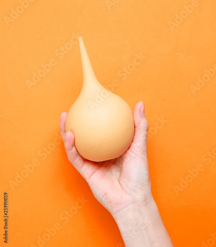 Female hand holds orange medical enema on orange background. Top view, minimalism. photo