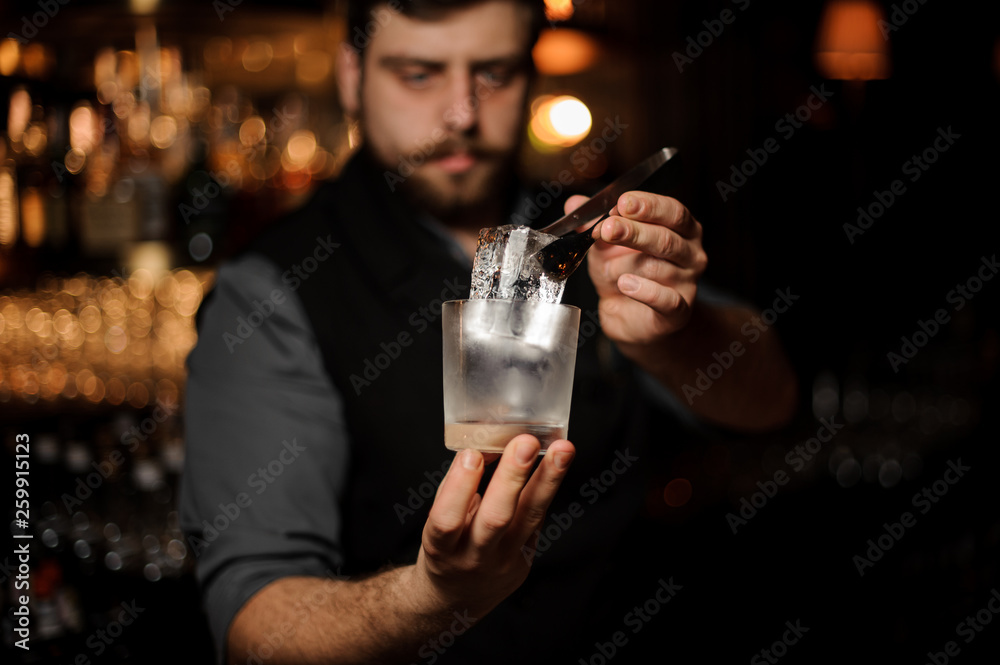 Bartender holding in hand an ice cube in tweezers putting it on the a cold matte cocktail glass