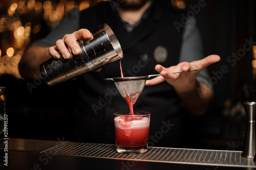 Professional bartender pouring a smooth pink cocktail through the sieve to the glass with one big ice cube