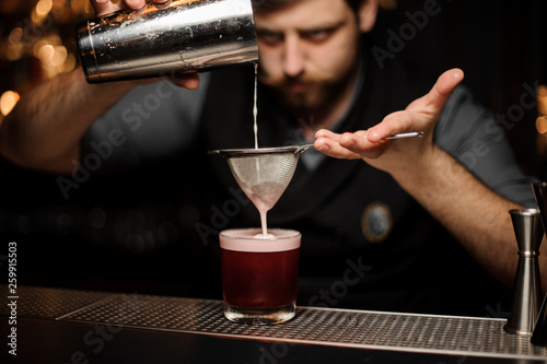 Bartender with a beard pouring a smooth crimson cocktail through the sieve to the glass with one big ice cube
