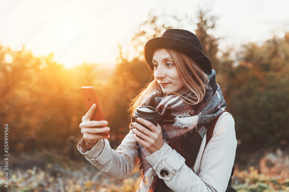 Woman drinking coffee in the autumn Park and using smartphone at the background of colorful sunset Woman drinking coffee in the autumn Park and using smartphone at the background of colorful sunset