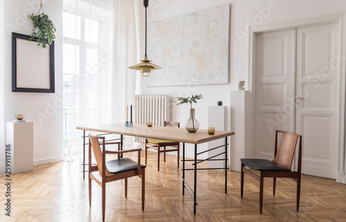 Stylish and modern dining room interior with design sharing table, chairs, gold pendant lamp, abstract paintings and elegant accessories. Tropical leafs in vase. Eclectic decor. Brown wooden parquet. © FollowTheFlow