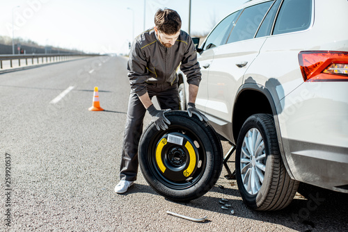Fototapet Road assistance worker in uniform changing car wheel on the highway