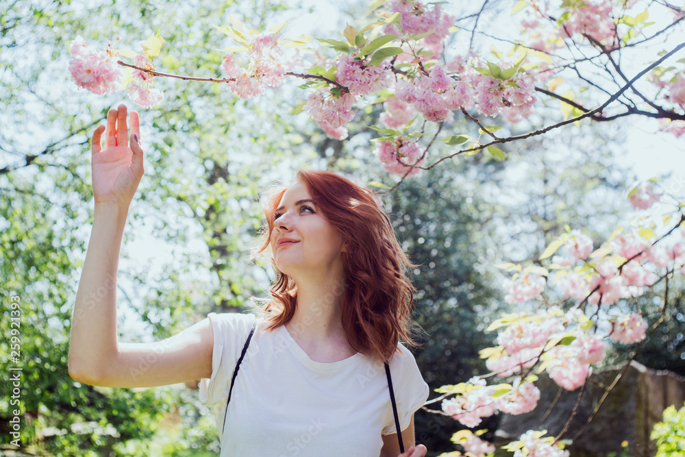 Young beautiful smilling redhead girl in white t-shirt in spring time blossom cherrytrees garden. Perfect time for a walk. Enjoy the spring and sunny day. Social media, beauty, blogger, vlogger. Young beautiful smilling redhead girl in white t-shirt in spring time blossom cherrytrees garden. Perfect time for a walk. Enjoy the spring and sunny day. Social media, beauty, blogger, vlogger.