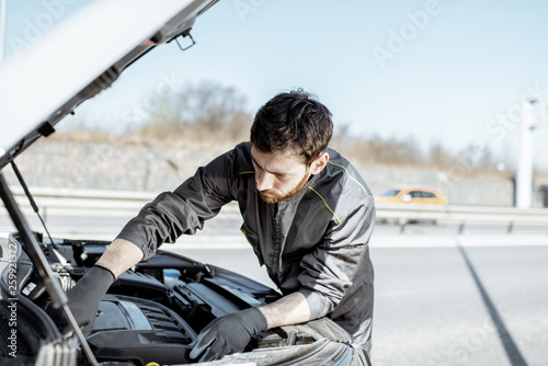 Handsome auto mechanic in uniform repairing engine of the broken car on the road