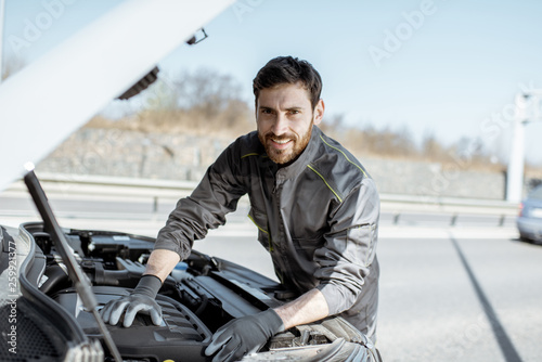 Portrait of a handsome auto mechanic or road assistance worker in uniform repairing engine of the broken car on the road © rh2010