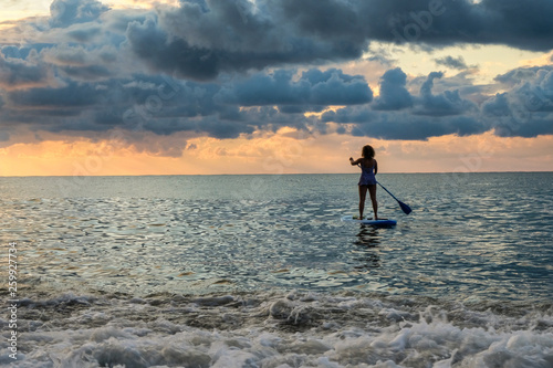 Young female paddling on SUP board in the sea at amazing dark sunset and waves. Sup surfing woman silhouette - active recreation in nature.  © Khrystyna Bohush