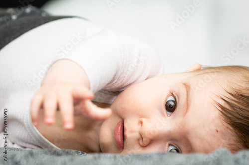 Adorable blond baby lying on his back on the white bed looking directly at the camera.