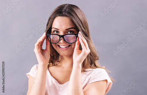Portrait of beautiful young woman wearing eyeglasses and smiling while standing isolated on gray background. Happy attractive girl holding glasses in hands. Woman putting on eyewear. Eyesight healt