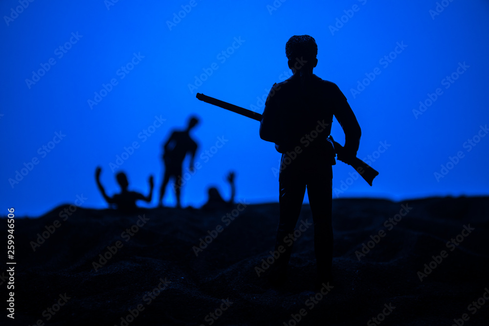 Obraz premium Man with riffle against zombie attack. Zombie apocalypse. Scary view of blurred zombies at cemetery and spooky cloudy sky with fog.